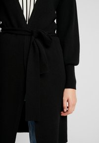 Svart stickad cardigan med ett knytbälte i midjan, med långa ärmar och en strukturerad tyg. Modellen håller tyget nära midjan.
