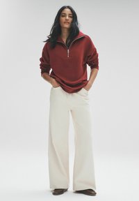 Sudadera roja con cremallera combinada con pantalones de pierna ancha en color crema. El material parece suave; la sudadera tiene un corte relajado, con mangas largas y un cuello profundo.