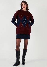 Burgundska oversized pulover s mornarsko modrim diamantom in belimi poudarki, kombiniran z mornarsko modro krilo in črnimi usnjenimi škornji do kolen.