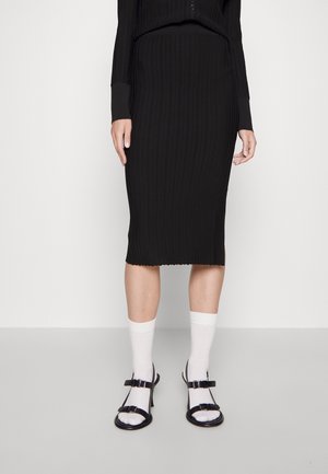 Kvinde iført en sort ribbet midi-nederdel, sorte sandaler med spænder og hvide crew-sokker mod en ensfarvet lys baggrund.