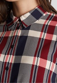 Primo piano di una persona che indossa una camicia a quadri rossa, blu navy, nera e bianca con colletto.