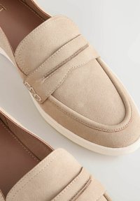 Beige suede loafers with a stitched detail, rubber sole, and a decorative strap across the front. Smooth texture and rounded toe design.