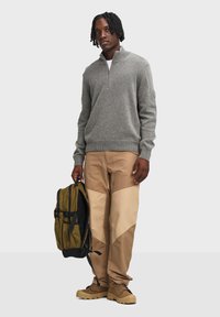 Szary sweter z dzianiny z zamkiem przy kołnierzu, beżowe spodnie cargo z blokowaniem kolorów oraz ciemnooliwkowy plecak. Beżowe obuwie dopełnia całość.