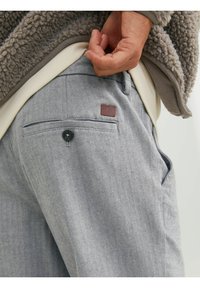 Pantaloni grigi a spina di pesce con una tasca posteriore dotata di bottone. Include un'etichetta in pelle. Sovrapposti a un esterno grigio testurizzato.