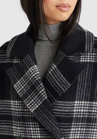 Femme portant un manteau à carreaux noir et blanc par-dessus un pull à col roulé gris côtelé, montrant le col et la partie supérieure de la poitrine.