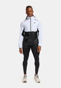 Hvid og sort vindjakke med lynlås; sorte leggings og lysegrå sneakers; har logo på brystet og kontrasterende paneler.