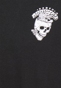 T-shirt noir avec un graphique blanc représentant un crâne avec des nouilles et des baguettes, portant les inscriptions "Accro aux ramen" et "100% Frais."