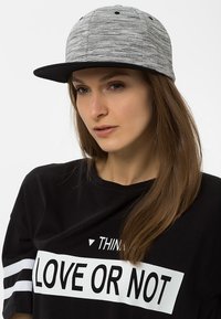 Sivá textúrovaná čiapka s čiernym okrajom, nosená s čiernym tričkom s bielym textom. Tkanina vyzerá hladko a neformálne.