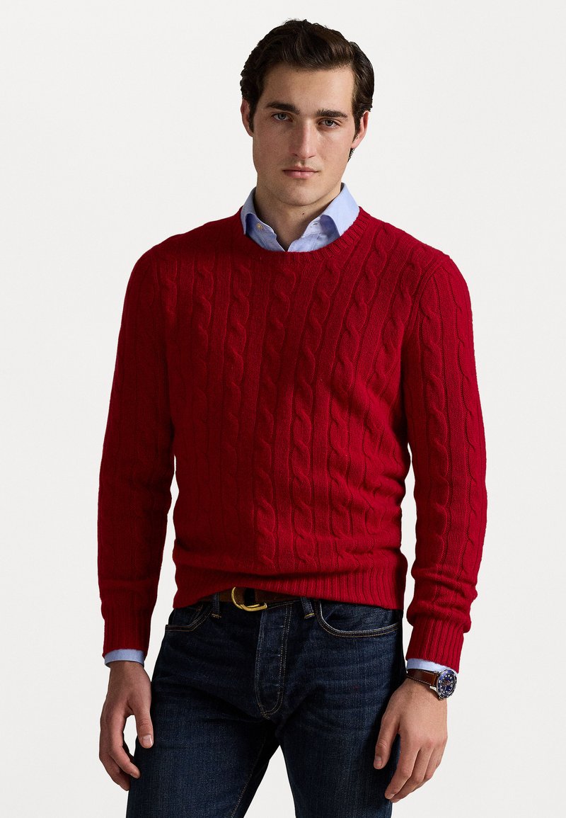 Suéter rojo de punto trenzado con cuello redondo, puños y dobladillo acanalados. Lleva puesto sobre una camisa de cuello azul claro, combinado con jeans de mezclilla oscura.