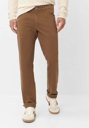 Mann trägt braune Freizeithose mit hochgekrempelten Bündchen und weiße Sneakers mit braunen Akzenten, steht vor einem einfachen hellen Hintergrund.