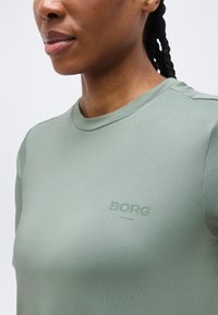 Γυναίκα που φοράει ανοιχτά πράσινο t-shirt με στρογγυλή λαιμόκοψη και το λογότυπο «BORG» στο στήθος, με πλεγμένα μαλλιά μαζεμένα προς τα πίσω, ουδέτερο φόντο.