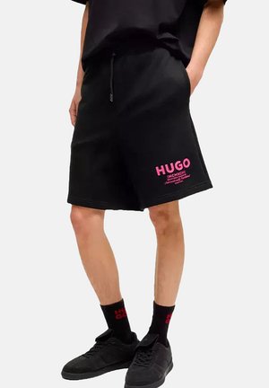 Svarta bomullsshorts med dragsko i midjan. Har rosa "HUGO"-text tryckt på vänster ben. Bärs tillsammans med svarta strumpor och skor.