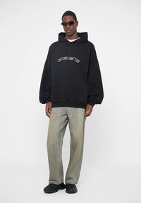 Schwarzer übergroßer Hoodie mit einem Reißverschluss vorne und weißem Logotext, kombiniert mit hellgrünen, weiten Hosen und schwarzen Sneakers. Accessoires umfassen Sonnenbrillen.