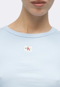 Φωτεινό γαλάζιο ριμπ T-shirt με στρογγυλή λαιμόκοψη και ετικέτα λογότυπου Calvin Klein με κόκκινα γράμματα σε λευκό φόντο.