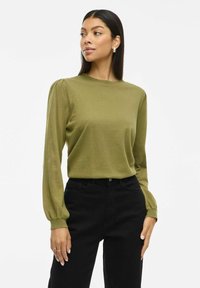Olijfgroene top met lange mouwen en een ronde hals, met opgezette schouders en een zachte textuur, gecombineerd met zwarte high-waisted broek.