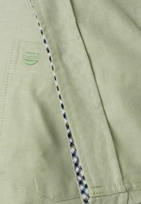 Lichtgroene stof met een gestructureerde afwerking, met een geruite patroon in blauw, wit en groen langs de naad en een circulair logo.