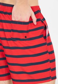 Shorts de bain rouges avec des rayures horizontales bleu marine, une taille élastique et une poche latérale avec un bouton à pression. Tissu doux et léger.