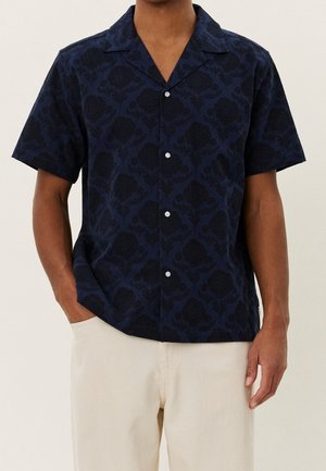 Homme portant une chemise bleu marine à manches courtes avec boutons, un motif floral sombre et un pantalon beige clair.