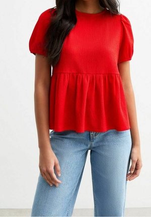 Mujer que lleva una blusa texturizada de manga corta en rojo brillante con cintura entallada y vuelo, combinada con vaqueros denim azul claro.