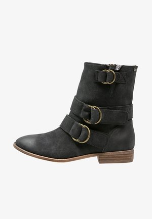 Black suede ankle boot with two wide straps, each featuring a circular gold-tone buckle. Low stacked heel and pointed toe design.