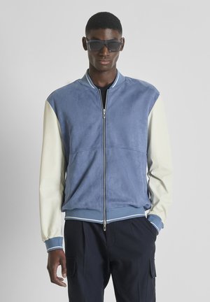 Hombre con gafas de sol oscuras de gran tamaño, chaqueta azul y blanca con cremallera y pantalones cargo negros, de pie frente a un fondo gris liso.