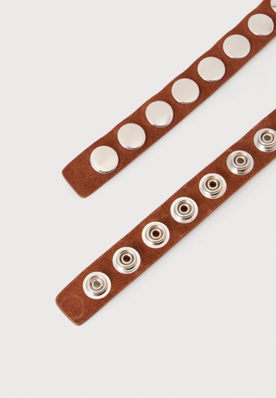 Deux sangles en daim marron, chacune dotée de six boutons-pression argentés ; l'une montre les têtes, l'autre les côtés douille, sur un fond uni.