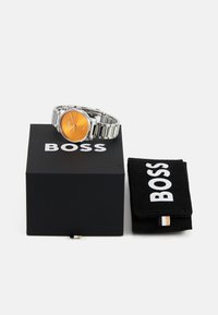 Reloj de acero inoxidable plateado con una esfera naranja, exhibido en una caja negra marcada "BOSS," junto a un estuche de ante negro.