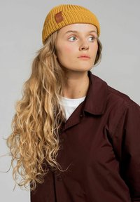 Musztardowo-żółta dzianinowa czapka z ryżowym splotem i małą brązową skórzaną łatką logo, noszona z ciemnoniebieską kurtką w kolorze burgundowym na białej koszuli.