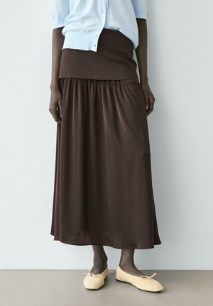 Donna che indossa una gonna midi marrone scuro arricciata, un cardigan azzurro chiaro con bottoni, calze scure e ballerine color crema, in piedi contro un muro semplice.