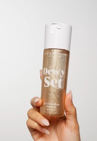 Gouden sprankelende settingspray in een doorzichtige fles met een witte dop. De tekst leest "Dewy Set" en "Setting Spray," met een soepele textuur.