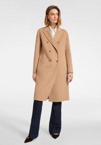 Mujer con abrigo largo beige de doble botonadura sobre camisa blanca y vaqueros azul oscuro, de pie con las manos relajadas a los lados, mirando hacia la derecha.