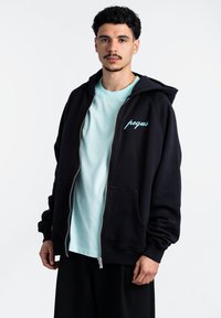 Juodas zip-up džemperis su erdvia putlia kepure ir priekinėmis kišenėmis, su akvamarino mėlyna logotipu ant krūtinės, sluoksniuotas virš šviesiai mėlynos spalvos marškinėlių.