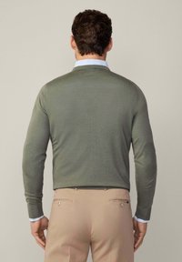 Pull olive green avec tricot texturé et coudières, associé à un pantalon beige clair. Présente un col bleu clair contrastant.