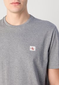 Vyras dėvi paprastą pilką trumpomis rankovėmis marškinėlį su mažu baltu lopu su raudona "CK" logotipu kairėje krūtinės pusėje.