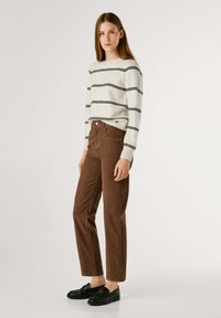 Gestreept sweater in crème met zwarte lijnen, gecombineerd met bruine corduroy broek. Model draagt zwarte instappers. Eenvoudige, casual styling.