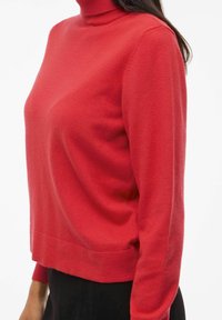 Crvena turtleneck dukserica od mekog materijala, s dugim rukavima i opuštenim krojem s ravnim rubom na dnu.