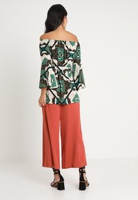 Donna che indossa un top a spalla scoperta con motivo e pantaloni larghi color corallo, in piedi con la schiena rivolta verso l'osservatore su uno sfondo semplice.