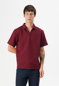 Burgundy short-sleeve polo shirt featuring a textured pattern and a half-open collar, paired with dark navy trousers.