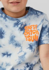 Blauwe en witte tie-dye T-shirt met korte mouwen en een geribbelde kraag. Heeft een oranje grafische tekst: "SURF'S UP CATCH THE SUN."