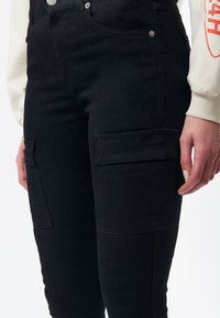 Czarne dżinsowe spodnie cargo z bocznymi kieszeniami, wysokim stanem i gładką fakturą. Widoczne są detale przeszycia oraz metalowe wykończenia.
