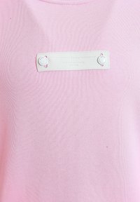 Top de punto rosa con una etiqueta blanca con texto en relieve, asegurada con botones decorativos. La tela tiene un acabado suave y texturizado.