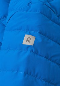 Giacca isolante blu con design trapuntato, tessuto leggero e un patch logo argento. Presenta cuciture visibili e una superficie liscia.