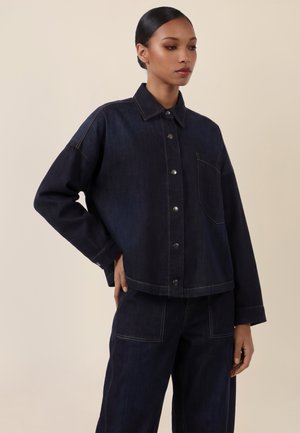 Frau trägt eine dunkelblaue Jeansjacke mit Druckknöpfen und passende weite Hosen, steht mit einer Hand in die Hüfte gestützt vor einem beigen Hintergrund.