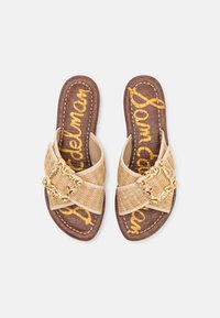 Sandales à glisser en paille tissée de couleur beige avec une boucle décorative en ton doré et une semelle intérieure en simili crocodile marron. Nom de la marque imprimé en jaune.