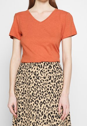 Frau trägt ein kurzärmeliges oranges V-Ausschnitt-Oberteil, das in einen hoch taillierten beigen Rock mit schwarzem Leopardenmuster und Knöpfen vorne gesteckt ist.