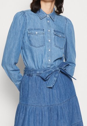 Niebieska sukienka z długim rękawem z denimu, z przodem zapinanym na guziki, kieszeniami na piersiach oraz pasem z tego samego materiału związanym w kokardę w talii.