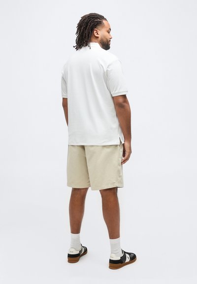 Άνδρας με ντραντλοκς στέκεται στραμμένος προς τα πίσω φορώντας λευκό πόλο, μπεζ σορτς, λευκές κάλτσες και ασπρόμαυρα αθλητικά παπούτσια.