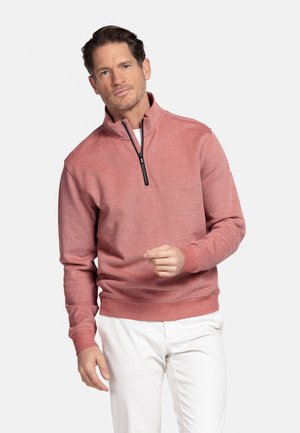 Man draagt een rozenkleurige trui met een kwart ritssluiting en witte broek, staand tegen een effen lichte achtergrond.