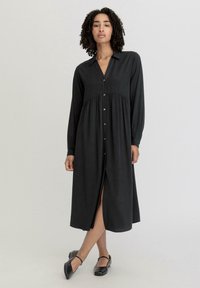 Schwarzes Langarmkleid mit durchgehender Knopfleiste, Kragen und Taillennaht. Es hat einen Schlitz vorne und eine glatte Stoffbeschreibung.