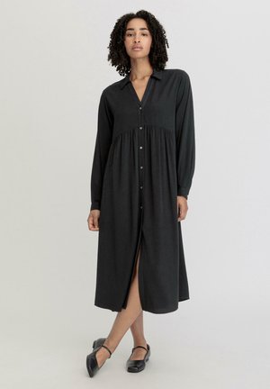 Zwart langemouwend jurk met een knoopsluiting aan de voorkant, kraag en taille-naad. Heeft een split aan de voorkant en een zachte stofstructuur.